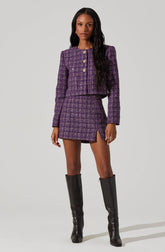 A woman is wearing a matching purple tweed jacket and mini skirt. The jacket features a textured pattern, a round neckline, and four decorative buttons down the front. The skirt has a similar textured pattern with a small slit on the side. She is also wearing tall black leather boots that reach above the knees.